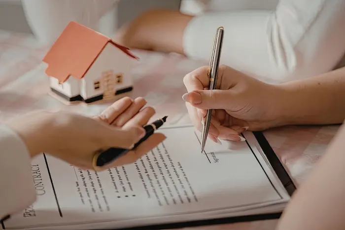 Deux personnes signent un contrat immobilier avec un modèle de maison sur la table, illustrant l’achat d’un appartement sécurisé.
