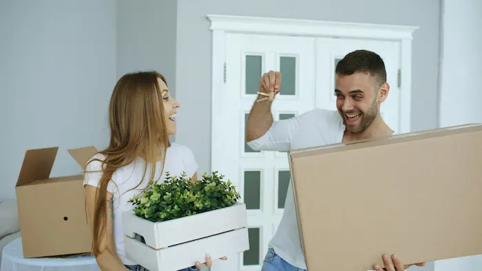 Couple portant cartons et clés devant porte vitrée, image illustrant l’achat immobilier à deux et le début d’une nouvelle vie commune. 