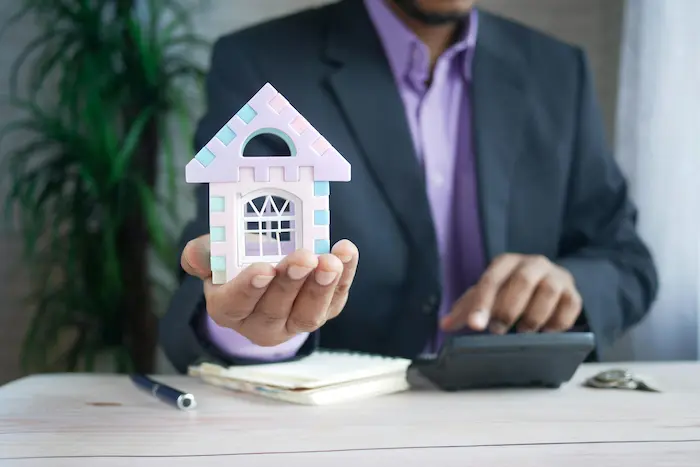 Personne en costume tenant une maison miniature colorée, image symbolisant la hausse de la taxe foncière et ses impacts sur les propriétaires.