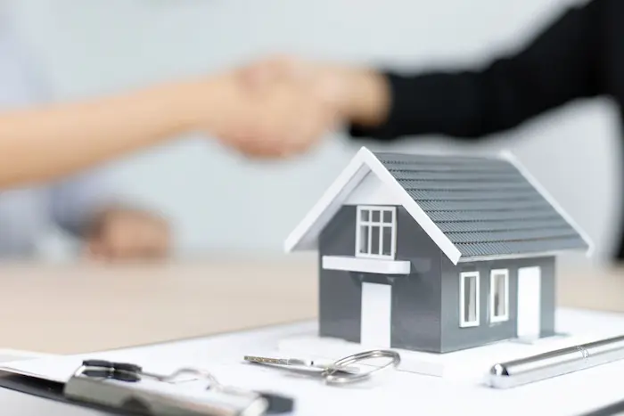 Maquette de maison avec clés et stylo sur document tandis que deux personnes se serrent la main illustrant la signature d’un contrat VEFA.