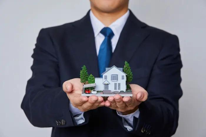 Main tenant une maison miniature bleue devant billets et pièces, illustrant le financement immobilier sans apport et l’accès facilité à la propriété.