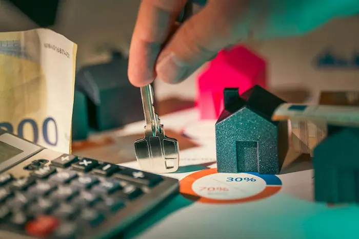 Main tenant des clés devant maquettes de maisons avec billet de 500 euros et calculatrice, illustrant le financement immobilier neuf.