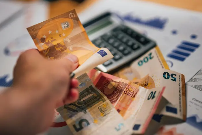 Main tenant des billets en euros avec calculatrice et documents financiers, illustrant les frais de notaire lors d’un achat immobilier.
