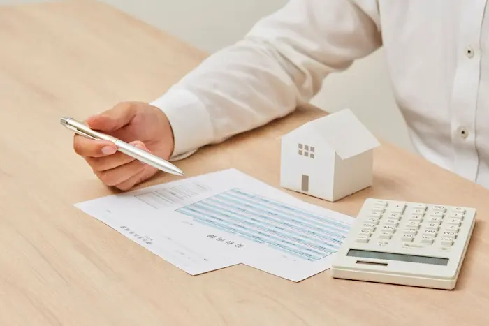 Personne en chemise blanche calcule avec maison miniature et documents financiers illustrant la recherche du meilleur taux pour un prêt immobilier neuf.
