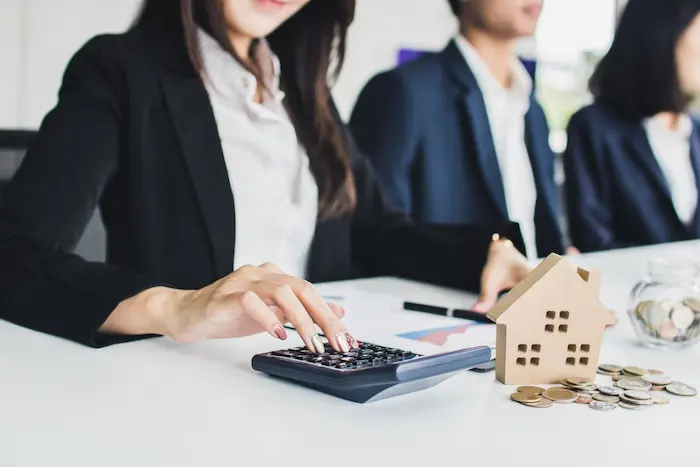 Personne calculant finances avec maison miniature et pièces, image illustrant le plafond PTZ pour l’achat immobilier et l’accès au prêt à taux zéro.