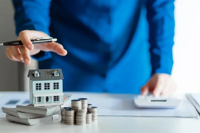 Personne en chemise bleue utilisant une calculatrice devant une maquette de maison posée sur des billets et des pièces, illustrant un simulateur de plus-value immobilière.