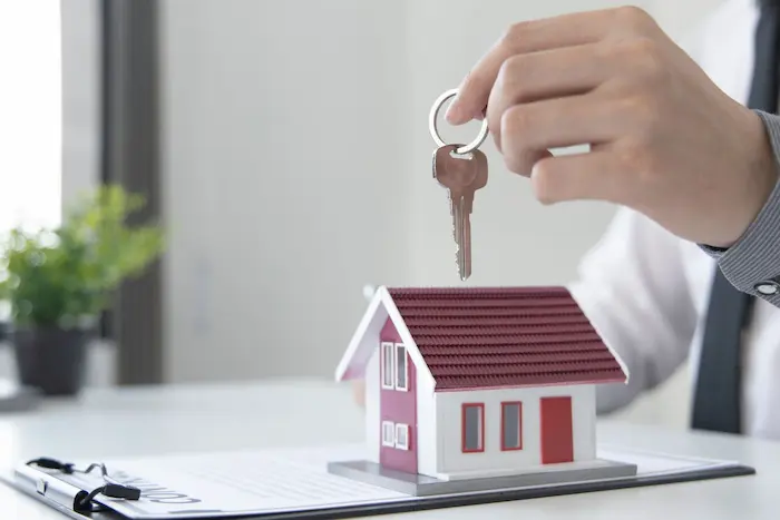 Main tenant une clé au-dessus d’une maison miniature sur documents, image représentant une simulation assurance prêt immobilier sécurisant capital et remboursement.