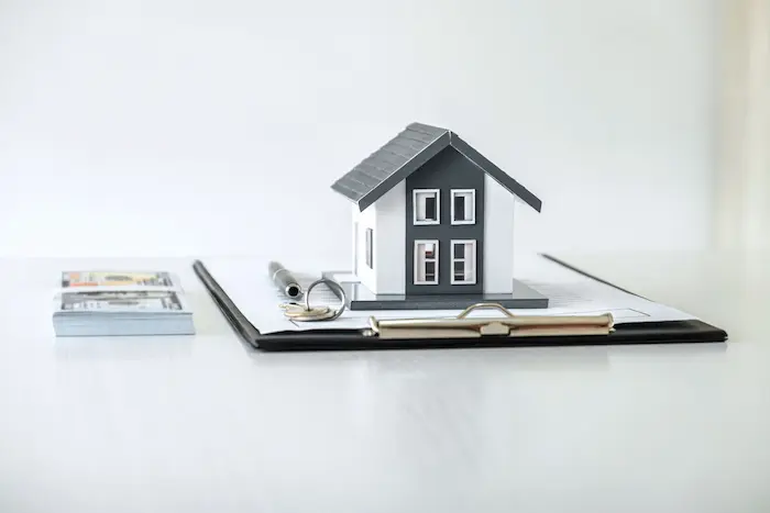 Maquette de maison posée sur un dossier avec billets et clés, image évoquant l’avantage fiscal de la TVA réduite à 5.5 pour l’achat d’un logement neuf.